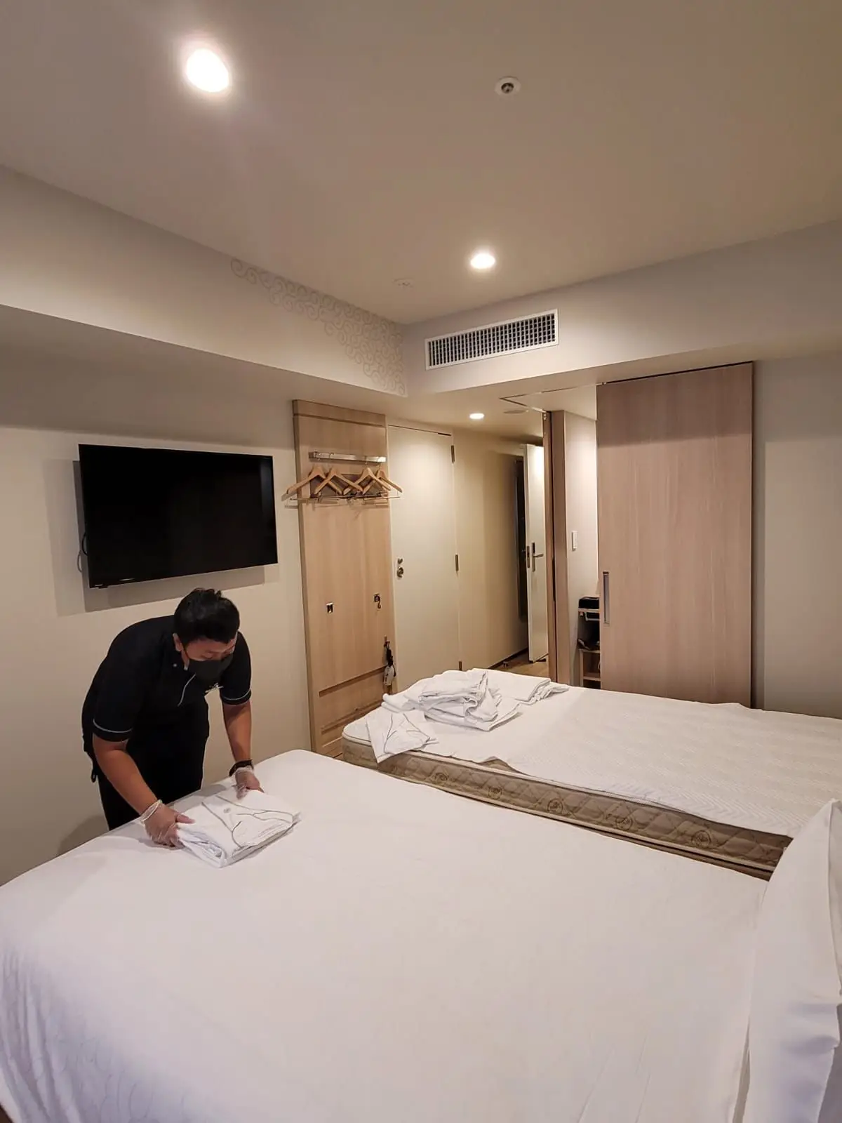 Person folding a towel on a neatly made bed in a well-lit hotel room.